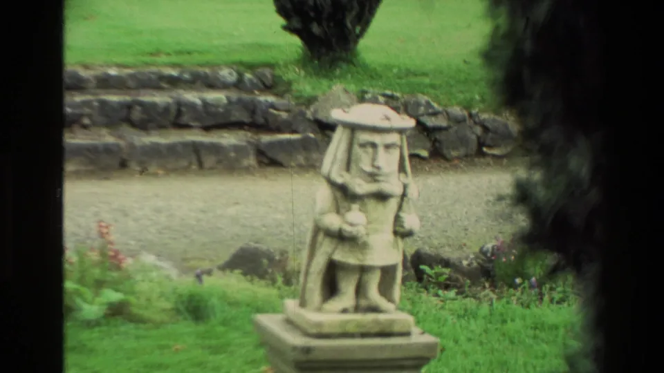 Close 1982 New Zealand travel frame of a stone garden figure on a pedestal with lawn, rock edging, and dark trees behind it.