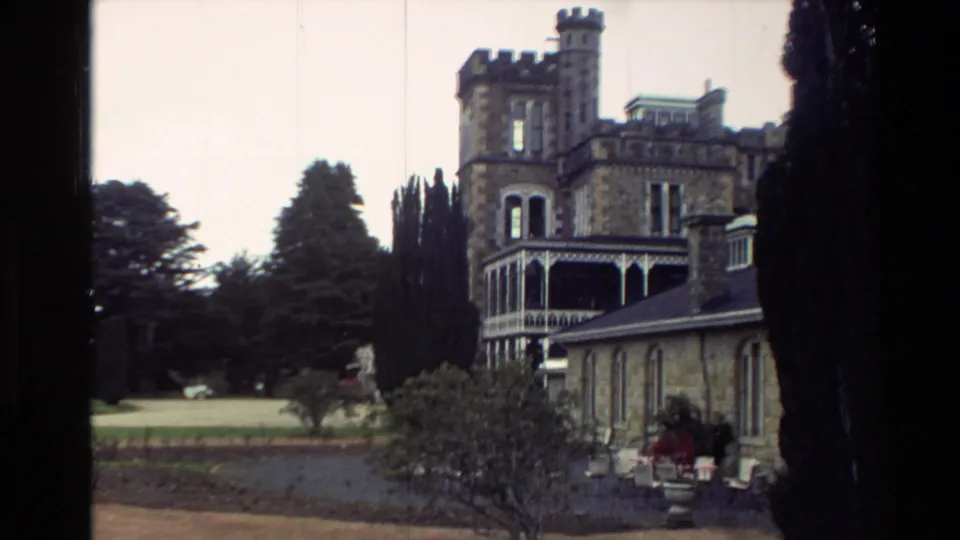 1982 New Zealand home-movie view of a castle-like stone building with turrets, veranda, trees, and garden foreground.
