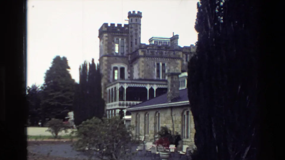 1982 New Zealand travel footage frame showing a turreted stone building and veranda framed by dark trees and garden planting.