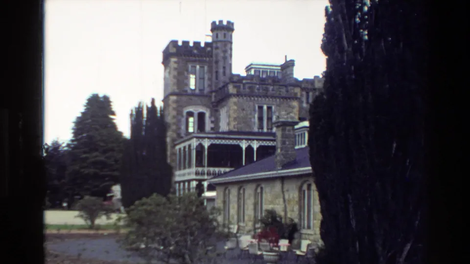 1982 New Zealand 8mm frame showing a castle-like stone exterior with crenellated towers, veranda details, trees, and garden beds.
