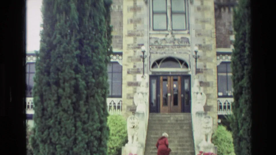 1982 New Zealand 8mm frame of a red-jacketed visitor ascending steps between stone lion statues toward a large arched doorway.