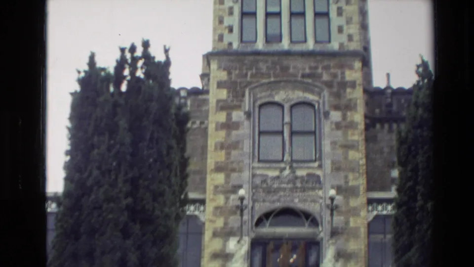 1982 New Zealand 8mm frame showing the castle-like entrance facade with arched doors, upper windows, stonework, and evergreen trees.