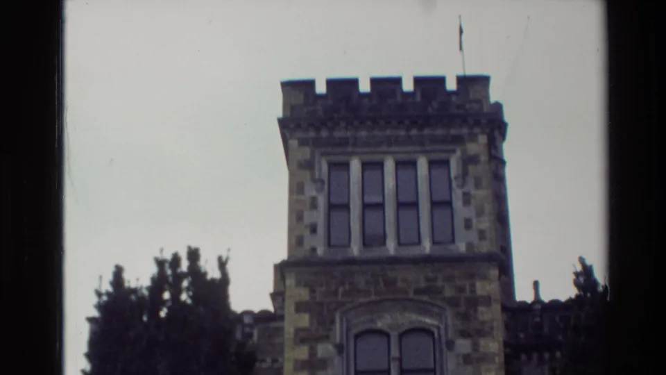 1982 New Zealand 8mm frame showing the upper tower, crenellated roofline, windows, flagpole, and trees of a castle-like stone building.
