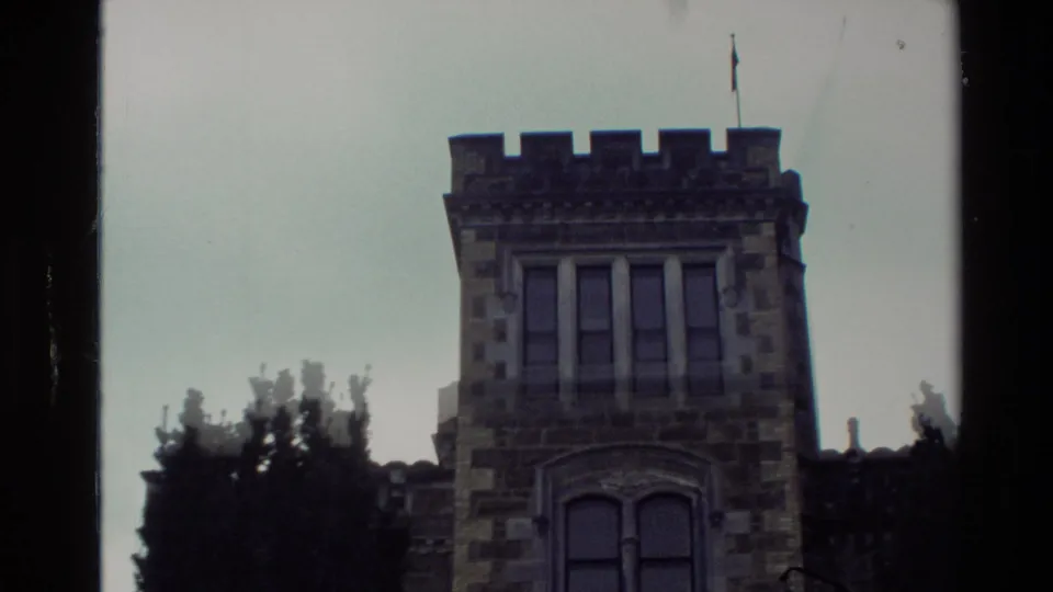 1982 New Zealand home-movie frame showing a stone castle-like building with arched windows, crenellations, trees, and a flagpole.