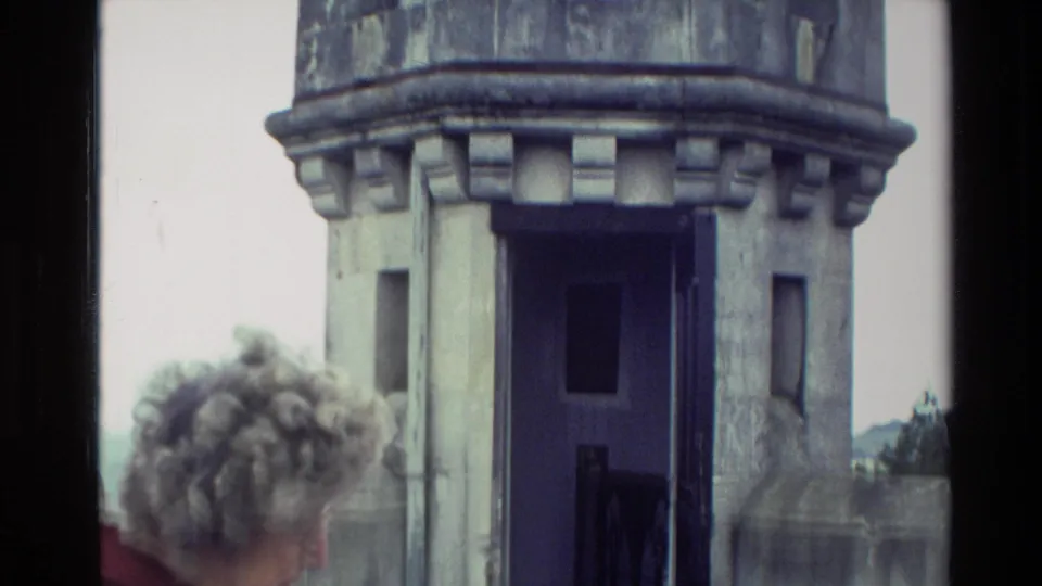 Elderly gray-haired woman in a red jacket near a round stone turret, filmed in New Zealand in 1982 home-movie footage.
