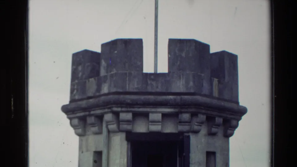 Close 8mm view of a round stone turret in New Zealand, with crenellations, flagpole, weathered stone, and narrow windows.