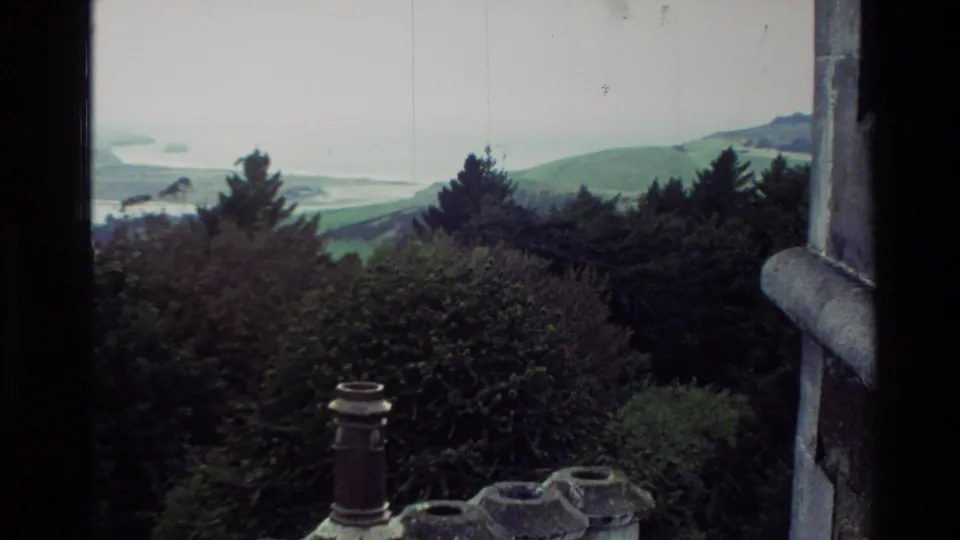 New Zealand home-movie view from near a stone tower toward trees, green hills, and a distant coastline in muted 8mm color.