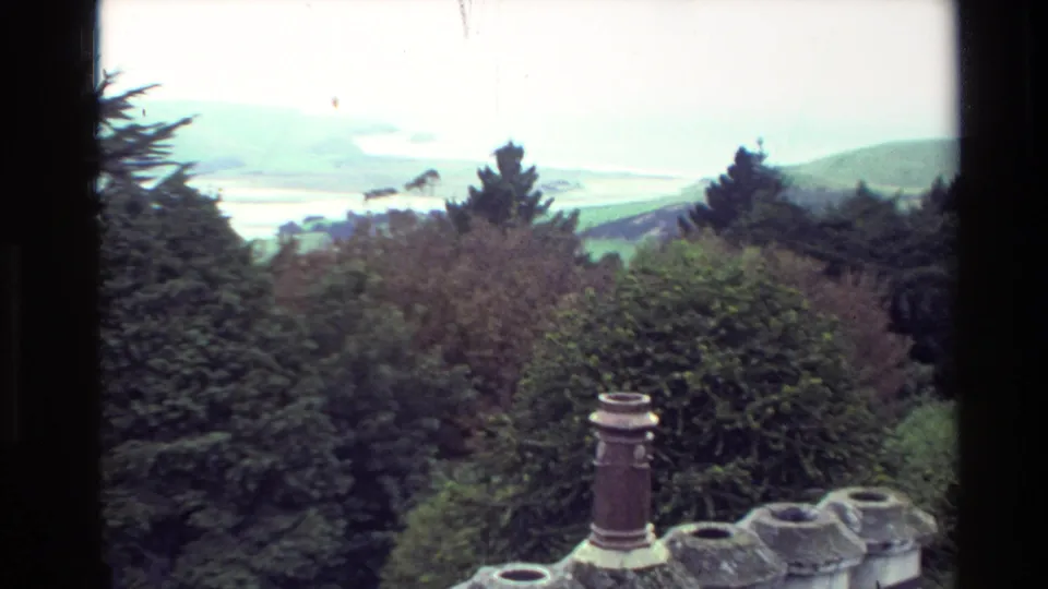 1982 New Zealand home-movie frame with shrubs, evergreens, chimney pots, hills, and a distant waterway under overcast light.