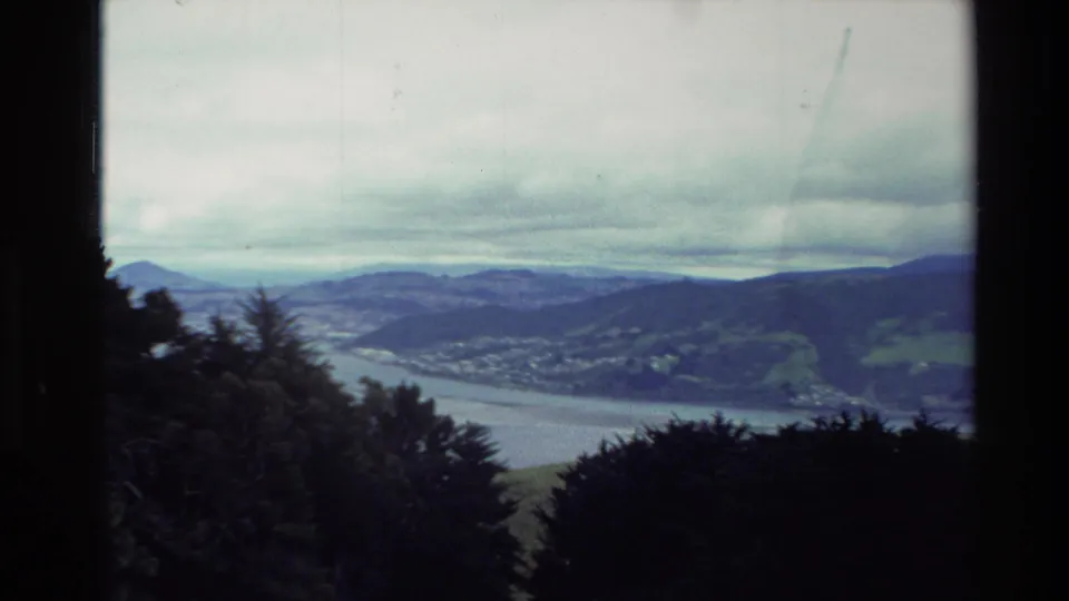 1982 New Zealand home-movie frame with a broader view across dark foreground trees, a wide waterway, layered hills, and low cloud.