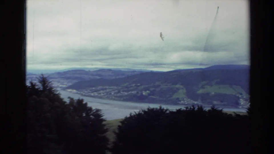 1982 New Zealand 8mm frame showing a river or bay, rolling hills, distant settlement, and dark foreground trees under a cloudy sky.