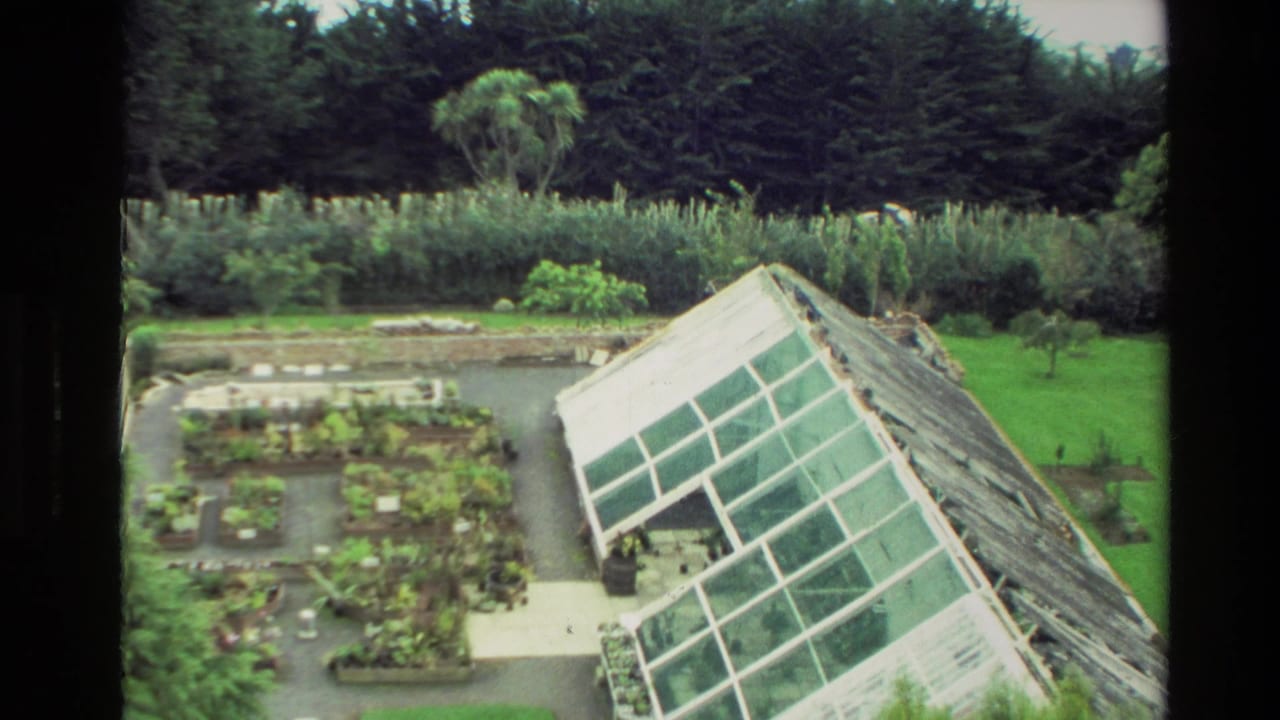Glass greenhouse beside garden plots with forest and lawn behind it.