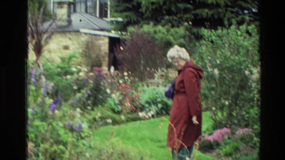 Elderly woman with white hair, red coat, and purple handbag walks through a New Zealand garden near a stone house exterior.
