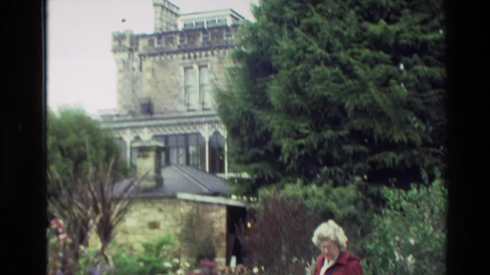 Garden and stone house exterior in New Zealand, with an elderly woman in red partly visible among trees and shrubs.