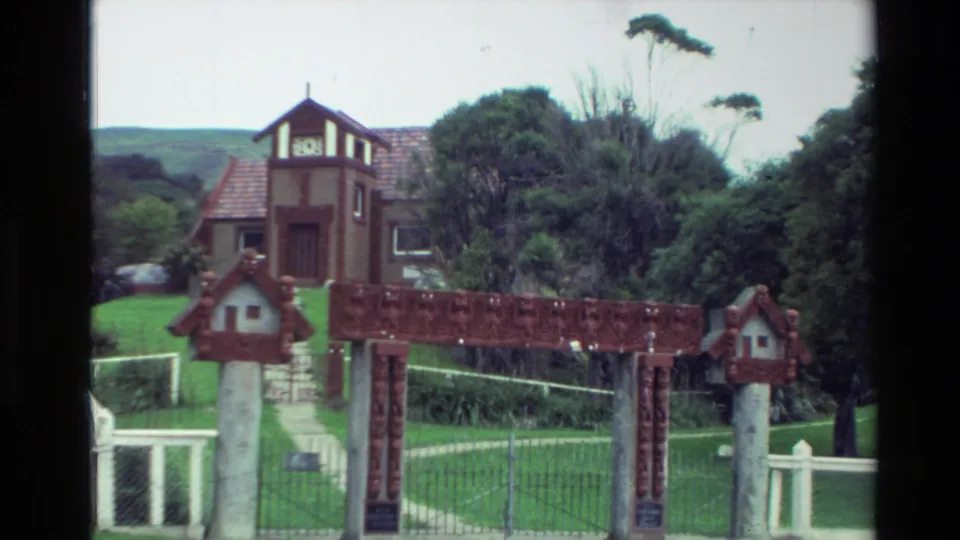 1982 New Zealand exterior view of a carved Māori-style wooden gate leading toward a towered house with lawn, trees, and hills behind.