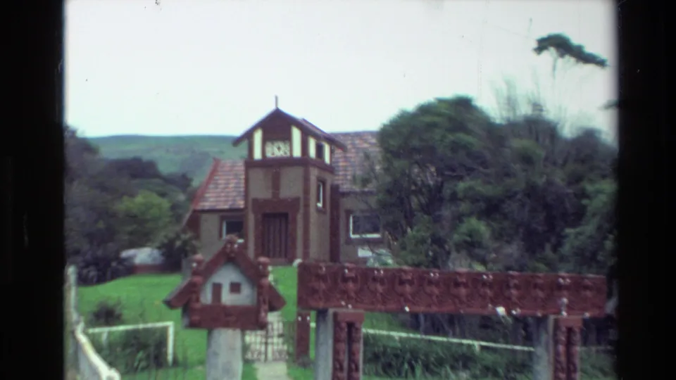 1982 New Zealand home-movie frame showing a towered house beyond a carved wooden entrance, with lawn, fencing, trees, and hillside background.