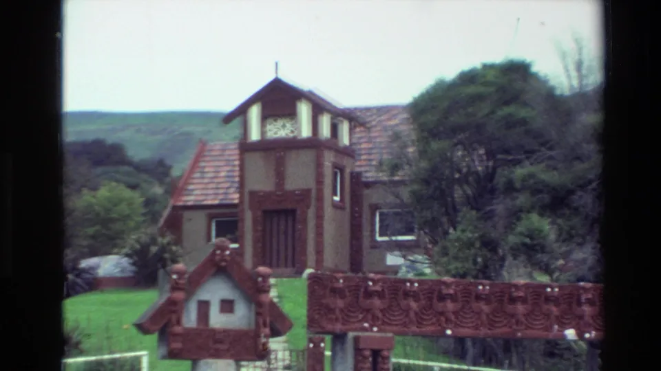 Closer 1982 New Zealand frame of the towered house and carved Māori-style gate, framed by greenery, white fencing, and low hills.