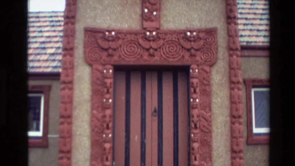 Close 8mm frame of a Māori meeting-house entrance with red carved posts, a vertical wooden door, and spiral lintel decoration.