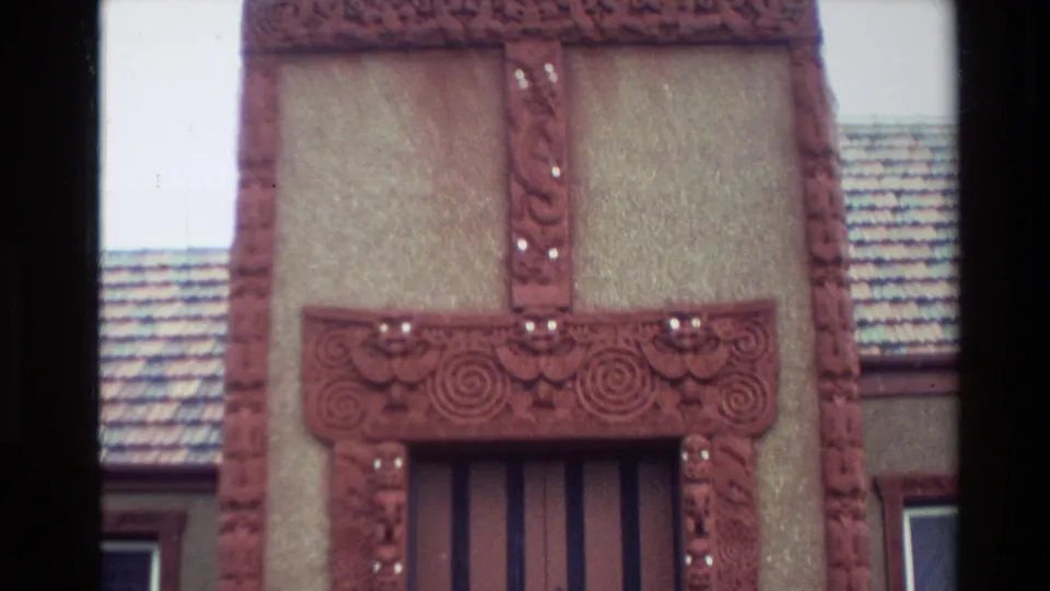 Home-movie close-up of red carved facade elements on a Māori meeting house, including posts, lintel spirals, roof tiles, and wall panels.