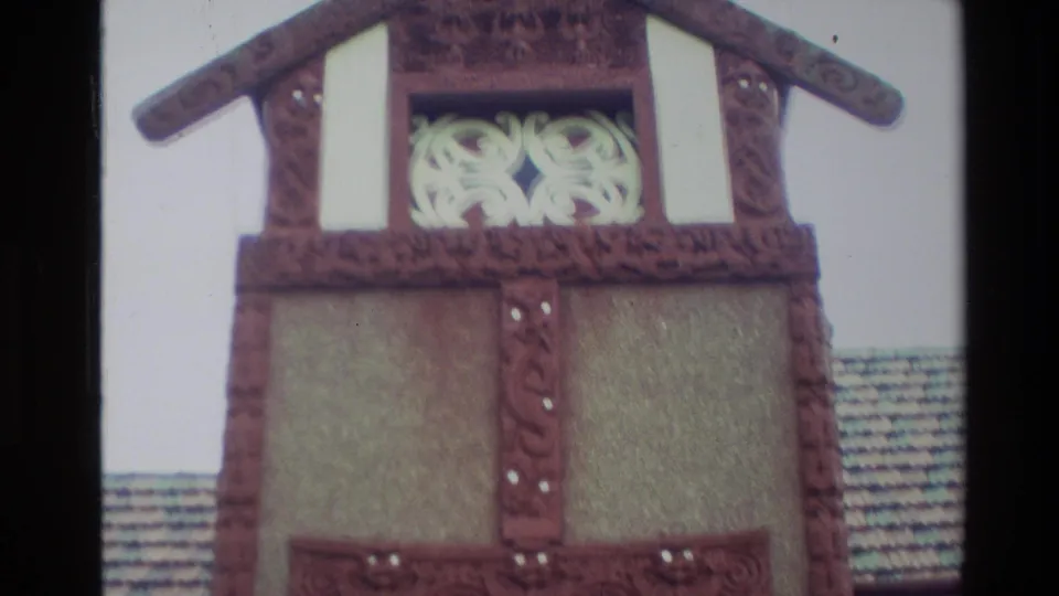 1982 New Zealand frame showing the upper gable of a Māori meeting house with carved red trim, pale decorative panel, and tiled roofline.