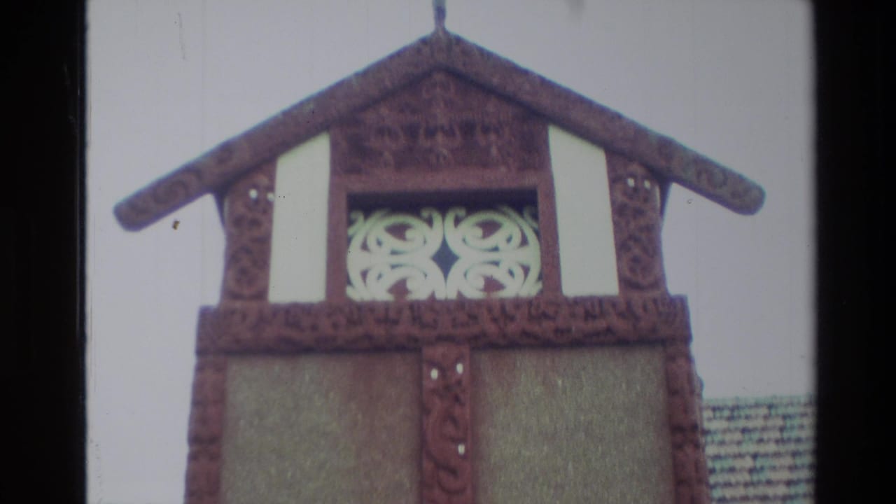 1982 New Zealand home-movie frame showing the upper front of a Māori meeting-house style façade with carved wood and white panel details.