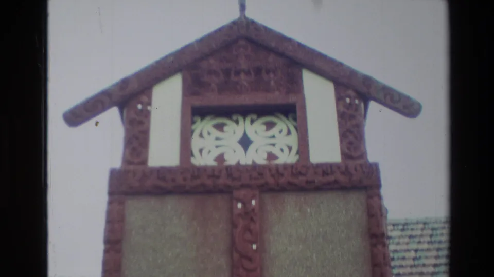 1982 New Zealand home-movie frame showing the upper front of a Māori meeting-house style façade with carved wood and white panel details.