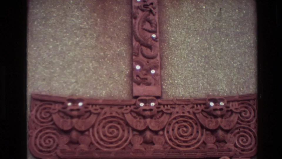 Close 1982 New Zealand home-movie view of carved meeting-house panels with spiral motifs, face forms, and white eye details.