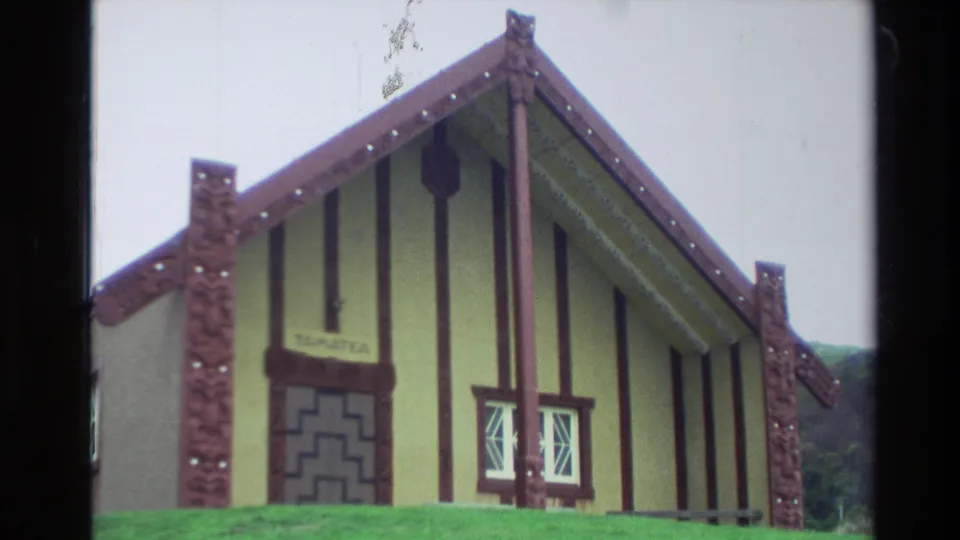 1982 New Zealand home-movie frame showing a gabled Māori meeting-house style exterior with carved posts, vertical trim, windows, and doorway.