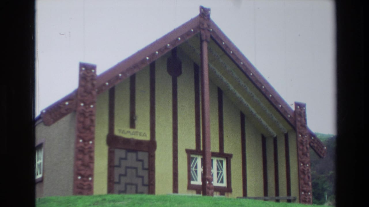 1982 New Zealand home-movie frame showing a carved meeting-house exterior with a visible Tamatea sign and decorative facade details.