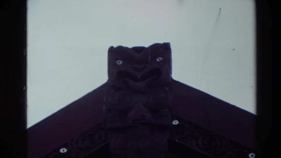 Close 8mm view of a dark carved roof figure on a New Zealand meeting-house exterior, with pale sky behind it.