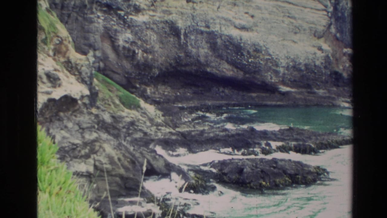 1982 New Zealand 8mm frame showing a rocky cove below layered coastal cliffs with grass, dark shoreline rocks, and turquoise water.