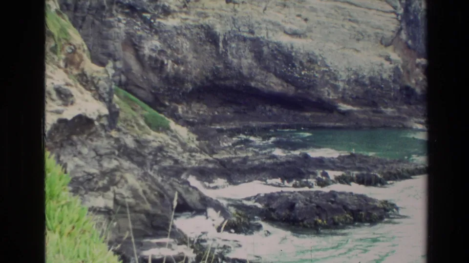 1982 New Zealand 8mm frame showing a rocky cove below layered coastal cliffs with grass, dark shoreline rocks, and turquoise water.