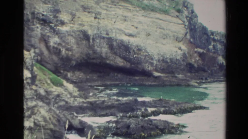 1982 New Zealand home-movie frame of stratified cliffs above a rocky shoreline with green-blue water and patches of surf.
