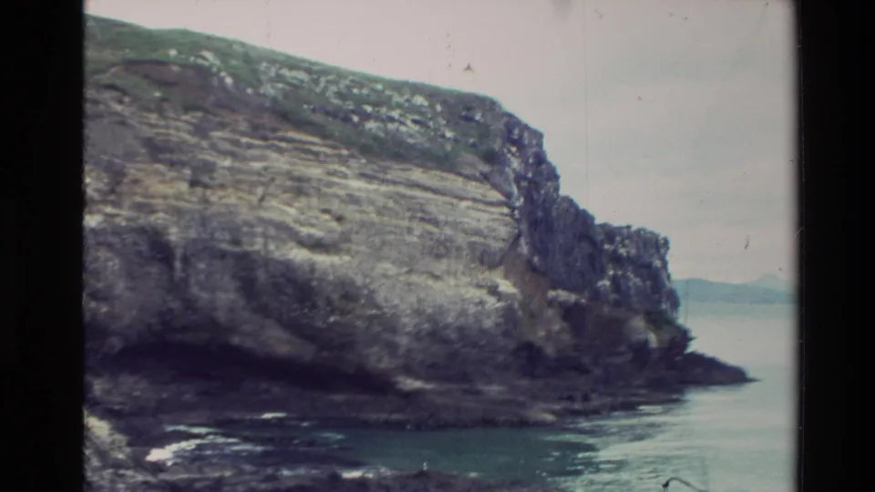 1982 New Zealand 8mm view of a broad layered cliff dropping toward calm water, with a faint distant shoreline beyond.