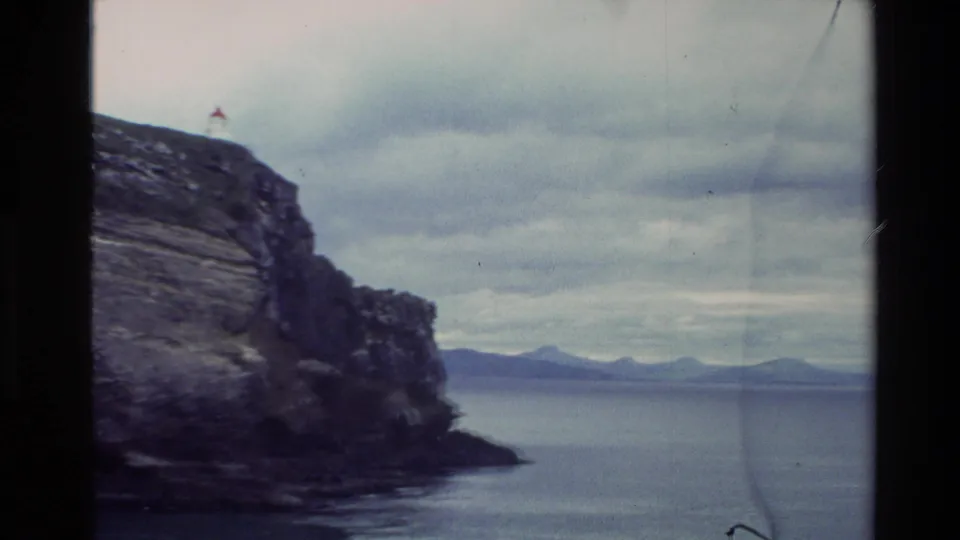 1982 New Zealand coastal frame showing a cliff edge, calm ocean, distant mountains, overcast sky, and a small lighthouse-like structure on the headland.