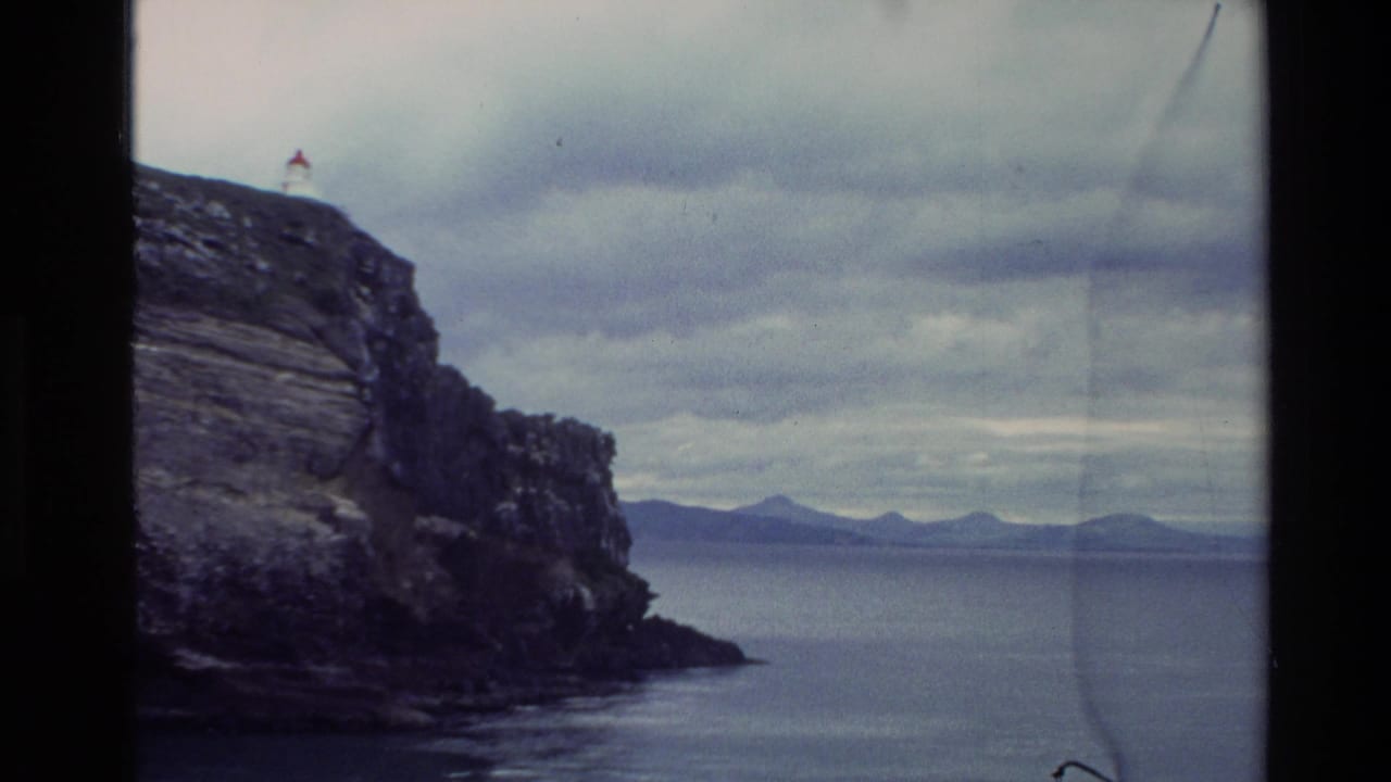 1982 New Zealand 8mm frame showing a dark sea cliff, distant horizon, cloudy sky, and ocean viewed from a coastal vantage point.