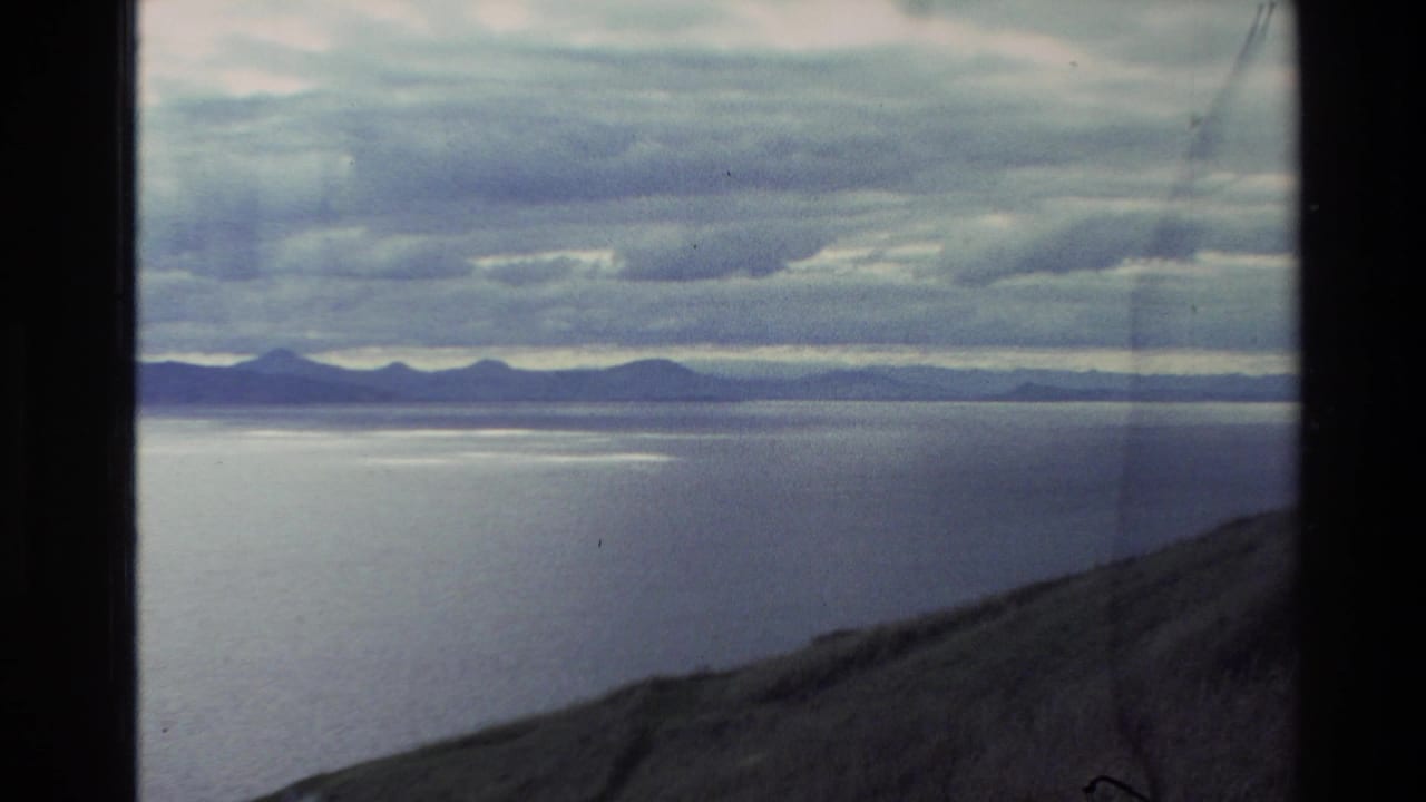 1982 New Zealand 8mm frame showing a broad lake or bay, distant mountains, cloudy sky, and sloped foreground land.