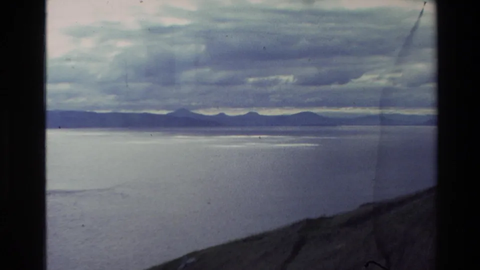 Wide 8mm view in New Zealand showing blue-gray water, far mountain silhouettes, layered clouds, and a sloping foreground bank.
