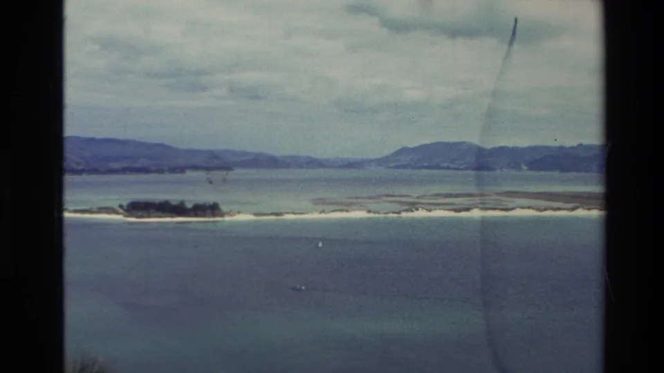 1982 New Zealand home-movie frame showing a small green island, pale sandbar, calm water, and distant shoreline hills.