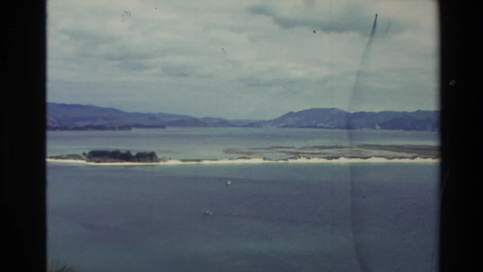 New Zealand 8mm frame from 1982 showing a low island, pale shoreline strip, blue-green water, and distant hills under cloud cover.