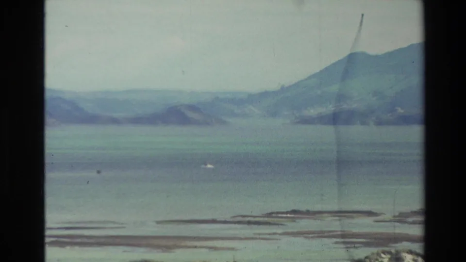 1982 New Zealand 8mm frame showing a calm blue bay, distant green hills, low island or sandbar forms, and cloudy sky.