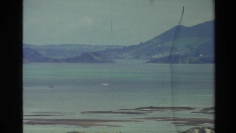 1982 New Zealand coastal view across still water toward distant hills, with low island forms and a small boat-like speck.