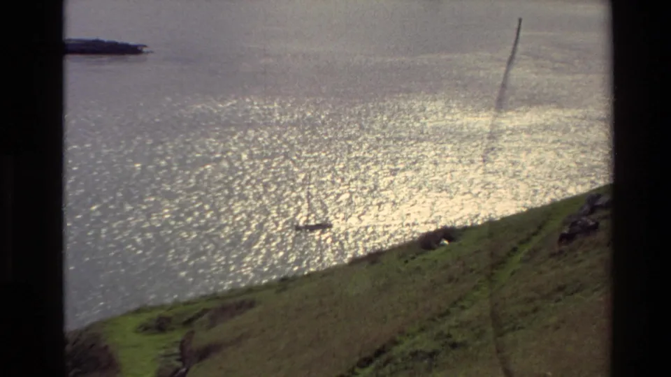 Wide 1982 New Zealand coastal frame with a grassy slope, bright ocean reflection, and rocky shoreline partly visible at the edge.