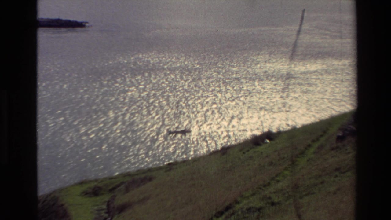 1982 New Zealand 8mm frame showing a small boat on bright rippled water below a grassy hillside and narrow path.