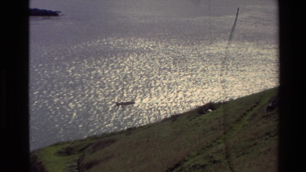 Sunlit ripples fill the water as a small boat passes below a sloping grassy bank.