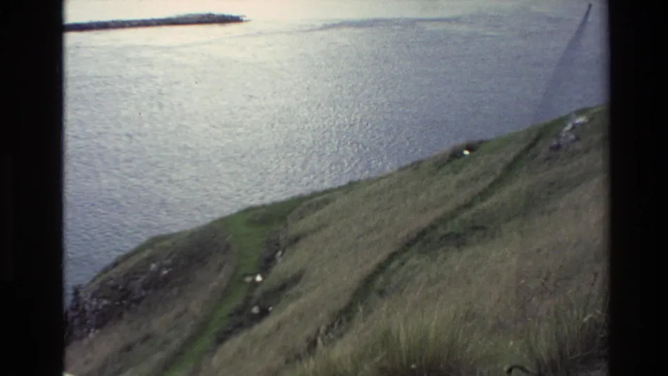 Soft 1982 New Zealand 8mm frame showing a green footpath on a hillside, with rocks, long grass, and water in the background.