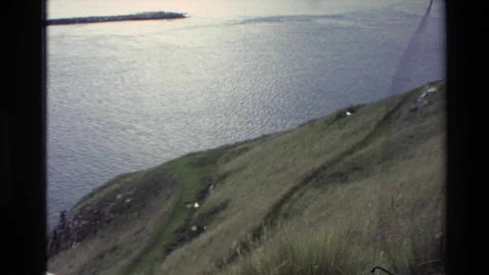 1982 New Zealand travel footage frame showing a sloped grassy headland, narrow path, scattered rocks, and calm coastal water.
