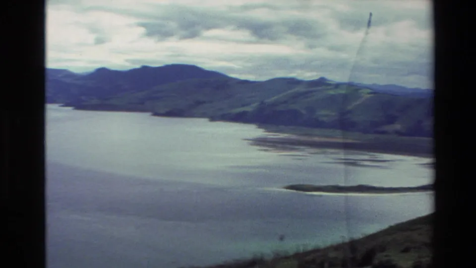 1982 New Zealand 8mm view of a wide bay, distant mountains, curved shoreline and sandbar beneath a cloudy sky.