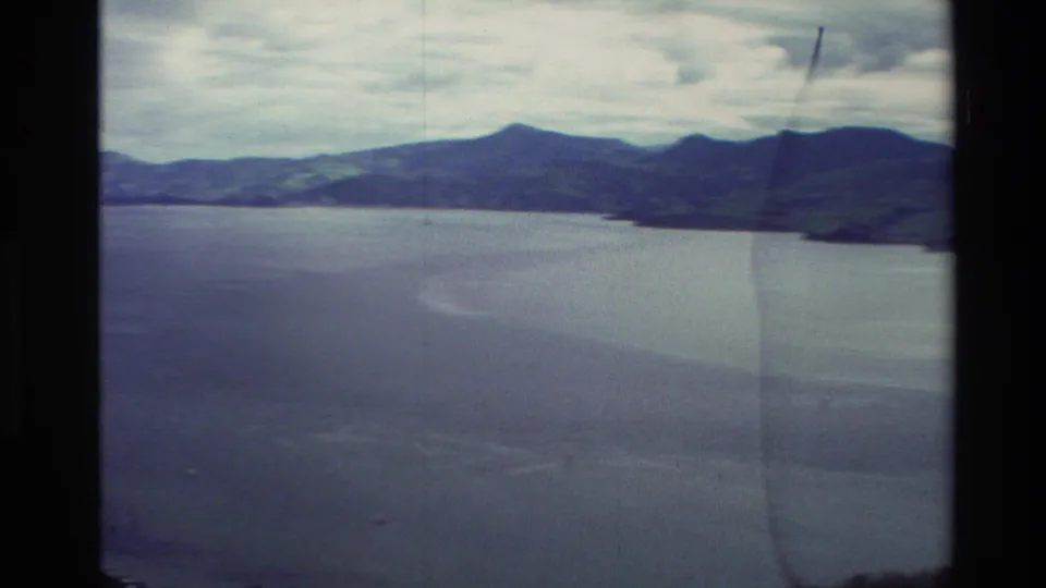 A New Zealand coastal panorama in 1982 8mm footage shows open water, dark mountains and a partly cloudy sky.