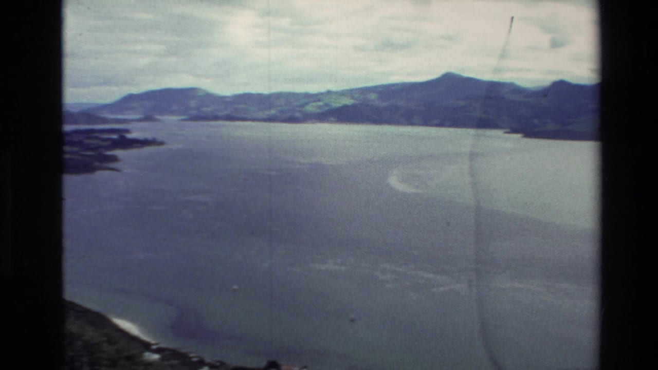 1982 New Zealand 8mm travel footage showing a broad lake beneath cloudy sky, with green hills and shoreline seen from an elevated viewpoint.