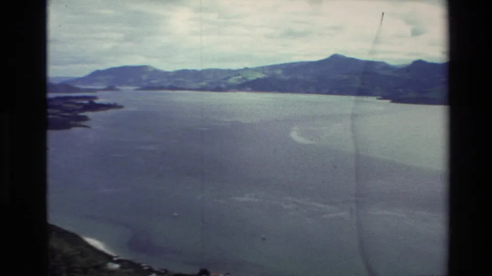 1982 New Zealand 8mm travel footage showing a broad lake beneath cloudy sky, with green hills and shoreline seen from an elevated viewpoint.